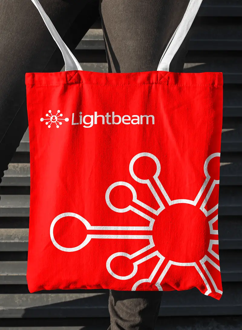 Red Lightbeam-branded tote bag with logo design, held by a person standing near a textured wall.”